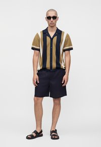 Homme portant des lunettes de soleil noires, une chemise à manches courtes boutonnée avec des rayures verticales marron, marine et blanches, un short marine et des sandales noires.