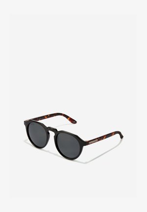 Sorte solbriller med runde mørke linser, ottekantet stel og arme med skildpadde-mønster, der har en lille logo-detalje.