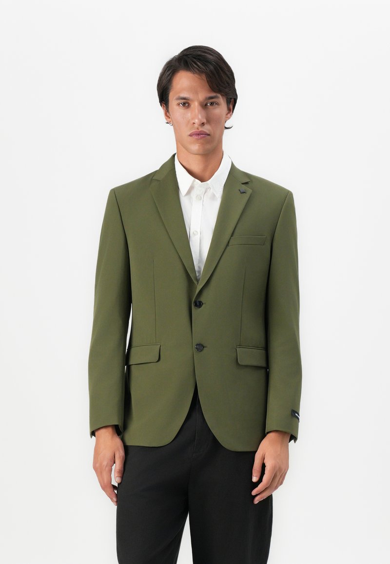 Man wearing olive green blazer over white buttoned shirt and black pants, standing against plain white background.