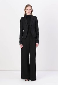 Černý střihaný blazer s jedním knoflíkem, ve spojení s přiléhavým černým rolákem a širokými černými kalhotami. Hladká textura, minimalistický design.