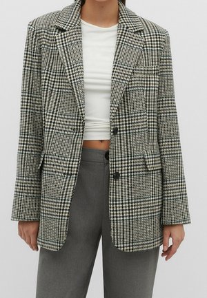 Femme portant un blazer pied-de-poule noir et blanc sur un crop top blanc et un pantalon taille haute gris, sur un fond uni.