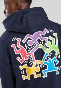 Hoodie bleu marine avec un graphique coloré au dos, représentant des figures stylisées en rouge, violet, vert, jaune et bleu. Matériau en coton.