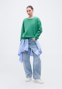 Sweat-shirt vert à encolure ronde, chemise rayée bleue et blanche nouée autour de la taille, jean en denim bleu clair, et baskets blanches.