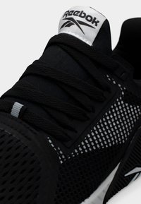 Κοντινή λήψη μαύρου αθλητικού παπουτσιού Reebok με δίχτυ στην πλευρική επένδυση, μαύρα κορδόνια και λευκό λογότυπο Reebok στη γλώσσα.