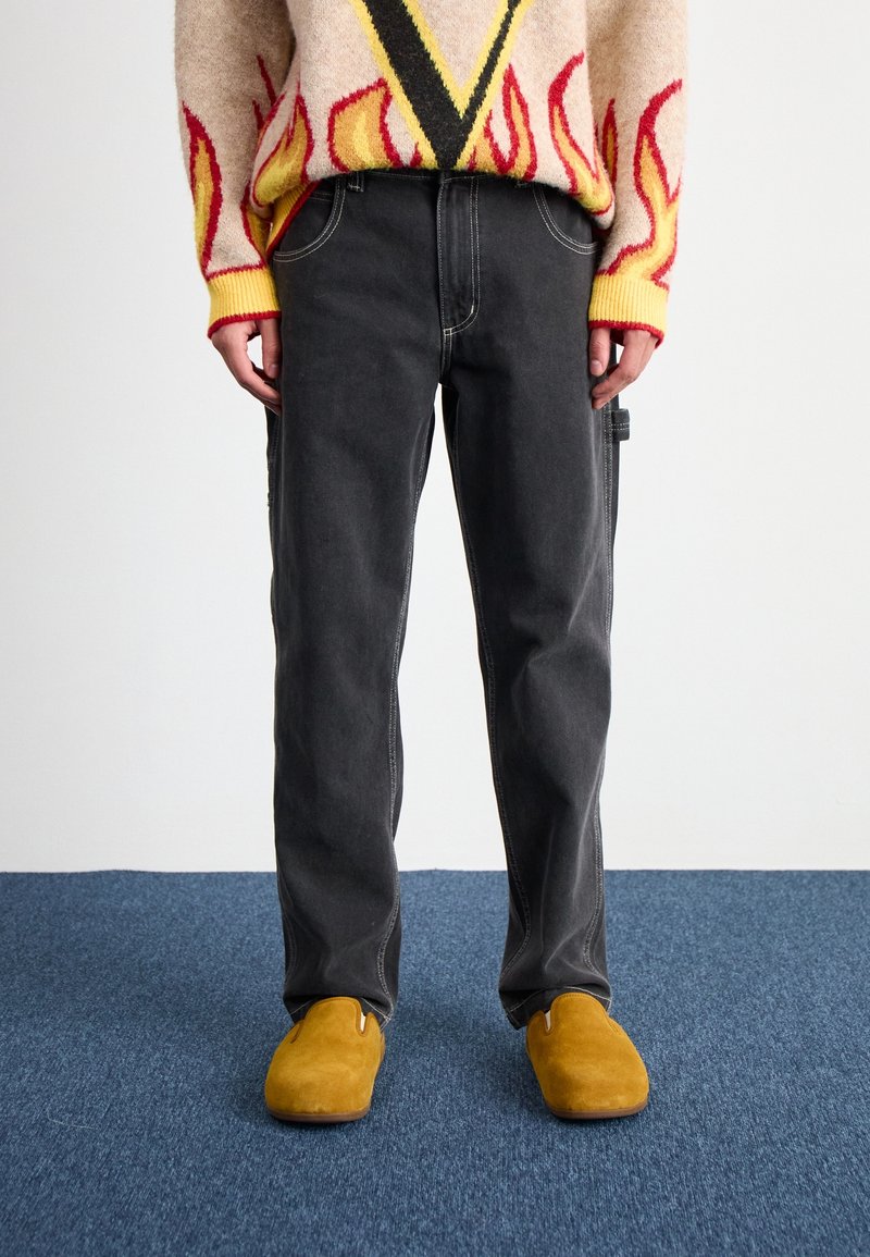 Personne portant un jean noir droit, un pull beige avec des motifs de flammes rouges et jaunes, et des chaussures à enfiler jaune moutarde sur un tapis bleu.