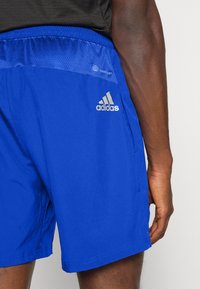 Siniset urheilushortsit, jotka on valmistettu kevyestä kankaasta; niissä on verkkokaulus ja hopeinen adidas-logo sivussa.
