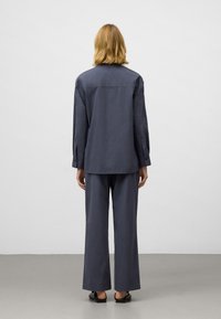 Person med axellångt blont hår som bär en lös, mörkgrå tröja med långa ärmar och vida byxor, stående mot en enfärgad vit bakgrund.