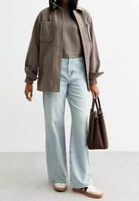 Surchemise grise oversize avec une poche, portée sur un pull gris côtelé, associée à un jean large bleu clair et un sac cabas marron.