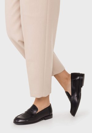 Person wearing beige cropped trousers and black leather loafers, standing with one leg crossed in front of the other on a plain background.