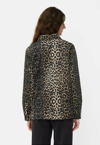 Frau mit langen braunen Haaren, die ein langärmliges Hemd mit Kragen in einem braun-schwarzen Leopardenmuster trägt und von hinten zu sehen ist.