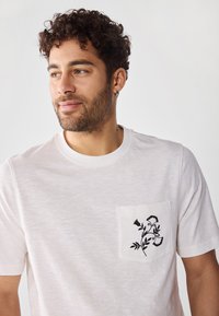 White cotton t-shirt featuring a chest pocket with a black floral design. Short sleeves and crew neck style with a textured fabric.