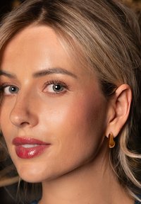 Goldfarbene Ohrringe mit einem geschwungenen, abstrakten Design; glatte Metalloberfläche; getragen von einem Model mit hellblonden Haaren und dezentem Makeup.