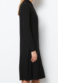 Kvinna iklädd en långärmad, knälång svart klänning med krage, visad i profil mot en ren vit bakgrund.