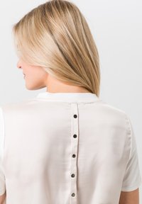 Korte mouwen top van lichtbeige stof met een gladde textuur, met een knoopsluiting langs de achterkant in zwarte accenten.