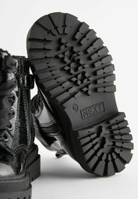 Bottes de combat noires brillantes avec des semelles épaisses et à motifs, fermeture éclair sur le côté, lacets, et la taille 6 marquée "NEXT" sous la semelle.