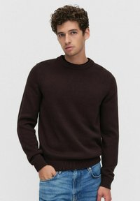 Donkerbruin gebreide trui met een ronde hals, geribde manchetten en zoom, gecombineerd met lichtblauwe jeans. Glad textuur en eenvoudig ontwerp.