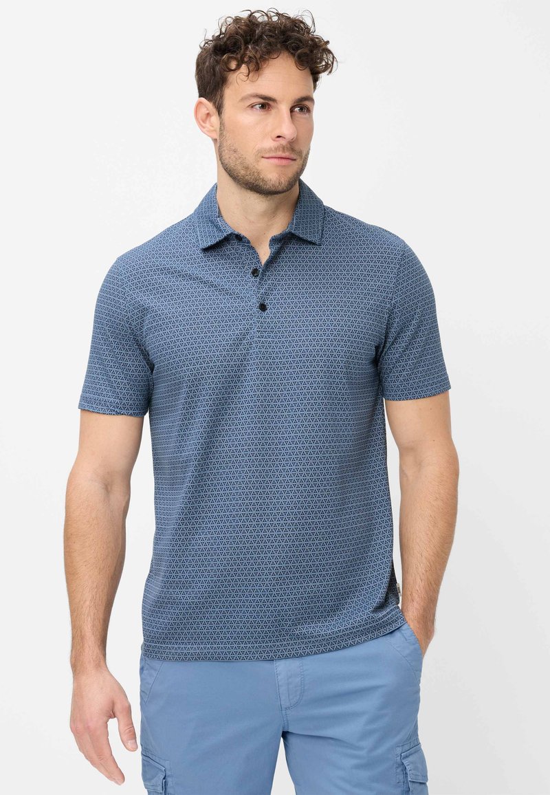 Homme aux cheveux bouclés portant un polo bleu à manches courtes à motif et un short cargo bleu clair, debout devant un fond blanc.