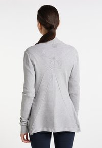 Woman with dark hair in a low ponytail wearing a light gray textured cardigan with geometric patterns on the back and dark pants.