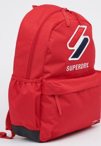 Rode rugzak met een gladde stoffen oppervlakte, voorzien van een wit en blauw geborduurd logo. Hij heeft een ritssluiting en een zwarte basis.