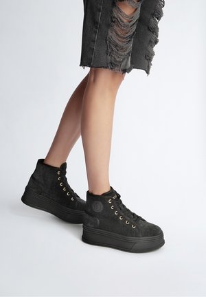 Legs wearing black high-top sneakers with thick soles and gold eyelets, paired with distressed black denim shorts against a plain background.