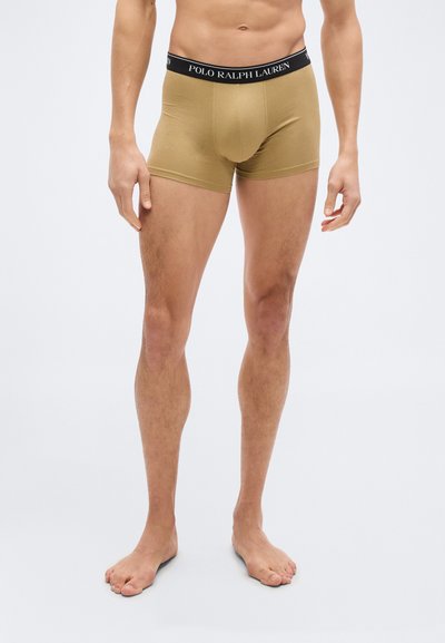 Άνδρας που φοράει μπεζ boxer briefs Polo Ralph Lauren, όρθιος ξυπόλυτος μπροστά σε απλό φωτεινό φόντο.