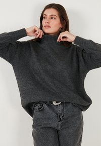 Donkergrijze oversized trui met een hoge nek, geribbelde manchetten en zachte textuur, gecombineerd met donkere jeans en een eenvoudige riem.
