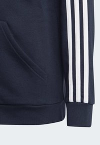 Sudadera marinera con un bolsillo frontal, dobladillo de canalé y tres rayas blancas en las mangas. El material parece suave y texturizado.