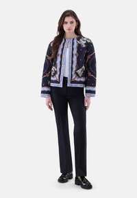 Gesteppten marineblauen Jacke mit blauem und goldenem Kettenmuster, kombiniert mit schwarzen Hosen und klobigen schwarzen Schuhen, mit einer lockeren Passform und Rundhalsausschnitt.