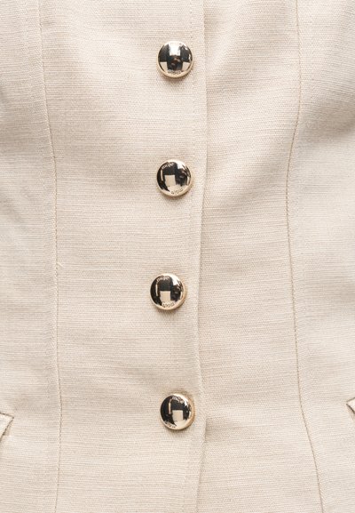 Κοντινό πλάνο μπεζ υφασμάτινης υφής σακακιού με τέσσερα λαμπερά στρογγυλά χρυσά κουμπιά τοποθετημένα σε κάθετη ευθυγράμμιση στο κέντρο.