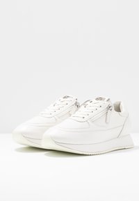 Valkoiset nahkakengät, joissa on teksturoitu viimeistely, vetoketjusuljin, valkoinen kumipohja ja sileät pintadetaljit.