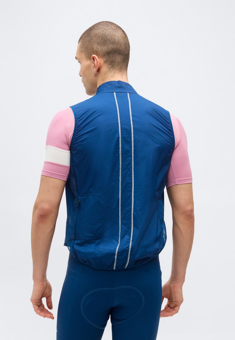 Man wearing a blue sleeveless vest over a pink short-sleeve top with white stripe, standing facing away against a plain background.