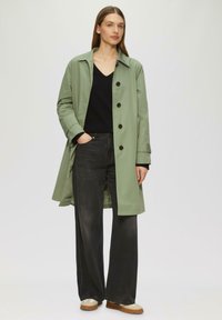 Lichtgroene trenchcoat met zwarte knopen, gelaagd over een zwarte trui en gecombineerd met donkere wijde jeans en beige schoenen.