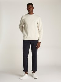 Sweatshirt creme com gola redonda e mangas longas, combinado com calças chino azul marinho e ténis brancos. Detalhe notável do logótipo no peito.