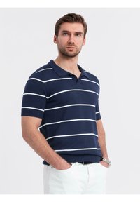 Námořnické modré polo tričko s krátkým rukávem a bílými vodorovnými pruhy, s výstřihem do V a žebrovanými manžetami, spárované se světle zbarvenými kalhotami.