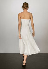 Femme aux cheveux attachés portant une robe blanche sans manches avec de fines bretelles marchant sur un sol sombre contre un mur clair uni.