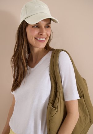 Usměvavá žena s béžovou čepicí s nápisem "OPTIMISMUS", bílým tričkem a velkou polstrovanou olivově zelenou taškou přes rameno na béžovém pozadí.