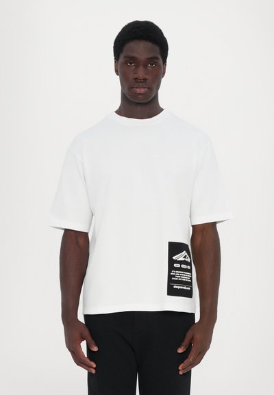 T-shirt bianca in cotone con maniche corte, vestibilità ampia e un'etichetta grafica nera sul lato sinistro inferiore con testo e logo.