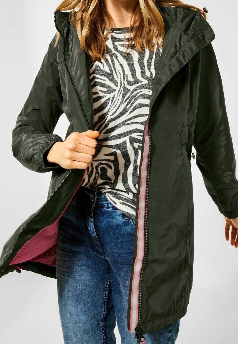 Olijfgroene waterdichte jas met capuchon, ritssluiting en roze voering. Gecombineerd met een grijs shirt met zebraprint en een blauwe spijkerbroek.