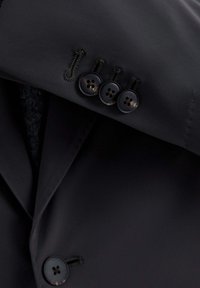 Chaqueta de traje negra con una solapa prominente, tres botones y costuras decorativas. Tejido suave con detalles de textura sutil.