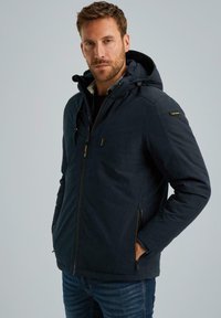 Giacca con zip blu navy dotata di cappuccio, tasche con zip e una texture liscia. La giacca presenta un design minimale con accenti di branding discreti.