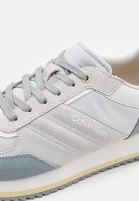 Lichtgrijze sneakers gemaakt van leer en suède met platte veters, een gewatteerde kraag, een logo en contrasterende blauwe accenten op de zool.