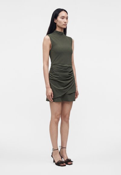 Femme debout en robe sans manches vert olive avec jupe froncée et sandales à talons noires sur un fond uni clair.