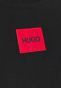 Černé tričko s červeným vyšitým čtvercovým nášivkem s nápisem "HUGO" v černé barvě, které poskytuje kontrastní designový prvek.