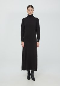 Abito midi nero a collo alto con maniche lunghe e spacchi laterali, realizzato in un morbido tessuto a maglia. Abbinato a stivali neri fino al ginocchio.