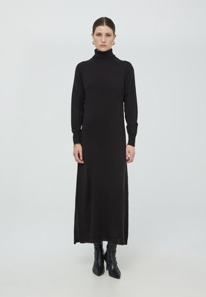 Abito midi nero a collo alto con maniche lunghe e spacchi laterali, realizzato in un morbido tessuto a maglia. Abbinato a stivali neri fino al ginocchio.