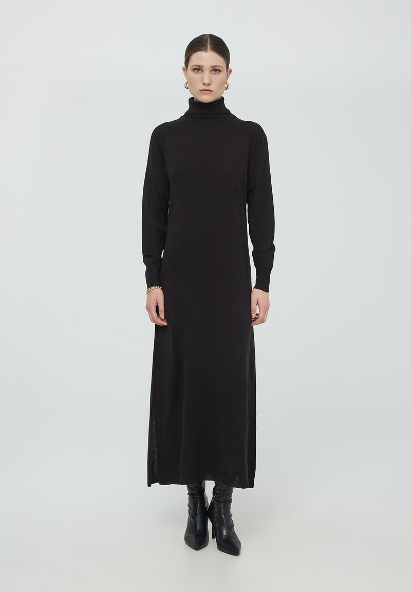 Abito midi nero a collo alto con maniche lunghe e spacchi laterali, realizzato in un morbido tessuto a maglia. Abbinato a stivali neri fino al ginocchio.
