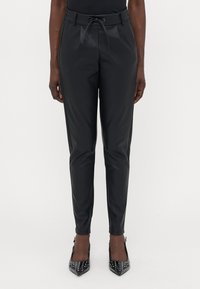 Pantalons noirs taille haute avec une ceinture à cordon, jambes fuselées et une texture brillante, associés à des escarpins noirs à talons aiguilles à clous.