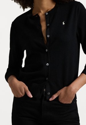 Osoba ubrana w czarny sweter rozpinany na guziki z białym logo Polo Ralph Lauren, ręce w kieszeniach, widziana od szyi do bioder.