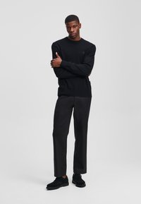Suéter negro con cuello redondo, elaborado en un tejido texturizado, combinado con pantalones negros de pierna recta y zapatos de ante negros. El modelo posa con los brazos cruzados.