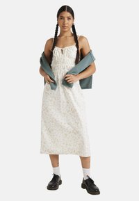 Vestido midi floral en blanco con patrones en tonos pasteles, cuerpo fruncido y tirantes tipo spaghetti. Un suéter verde azulado caído sobre los hombros. Zapatos negros.